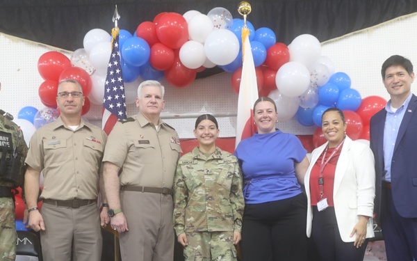 A group of seven individuals pose for a formal photo in front of a patriotic red, white, and blue balloon arch with an American flag. The group includes four military personnel in uniform and three civilians, suggesting an official military ceremony or event. The setting appears to be an indoor venue decorated for a patriotic occasion.