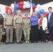 A group of seven individuals pose for a formal photo in front of a patriotic red, white, and blue balloon arch with an American flag. The group includes four military personnel in uniform and three civilians, suggesting an official military ceremony or event. The setting appears to be an indoor venue decorated for a patriotic occasion.
