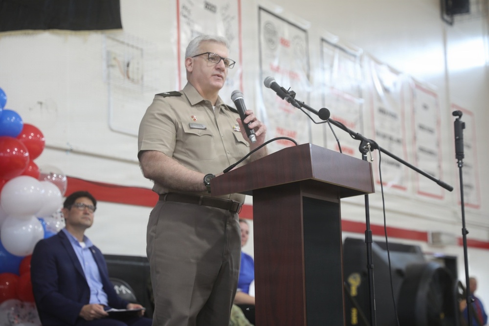 A military officer in tan/khaki uniform is delivering a speech at a podium while holding a microphone. The event appears to be a formal ceremony or official gathering, likely patriotic in nature given the red, white, and blue balloon decorations visible in the background. The setting appears to be an indoor gymnasium or assembly hall.