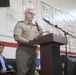 A military officer in tan/khaki uniform is delivering a speech at a podium while holding a microphone. The event appears to be a formal ceremony or official gathering, likely patriotic in nature given the red, white, and blue balloon decorations visible in the background. The setting appears to be an indoor gymnasium or assembly hall.