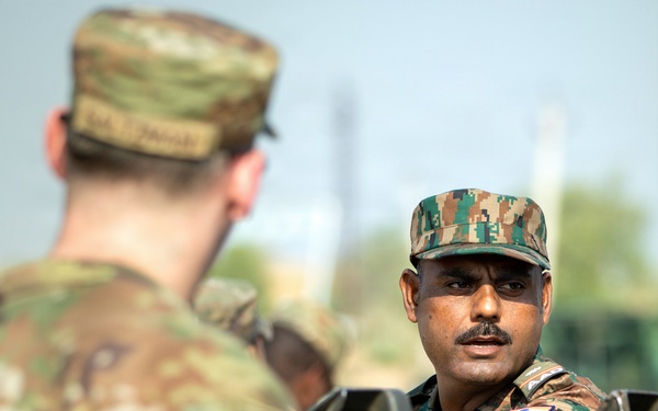 Two military personnel in different camouflage uniforms are engaged in conversation across what appears to be a military vehicle or equipment. The soldier in the foreground wears a MultiCam/OCP-pattern uniform, while the soldier in the background wears a digital camouflage pattern uniform with a visible flag patch consistent with Indian Army insignia. The image appears to document a joint military exercise or bilateral engagement.