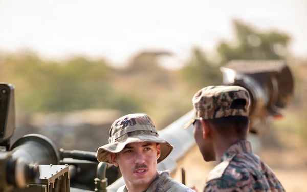 Two soldiers from different nations are engaged in a discussion next to what appears to be an artillery piece in an arid outdoor environment. The soldier on the left wears U.S. Army OCP (Operational Camouflage Pattern) uniform with a boonie hat, while the soldier on the right wears Indian Army camouflage with visible Indian flag and Army patches. This appears to be a joint military exercise or training exchange between U.S. and Indian armed forces.