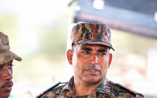 The image shows military personnel from at least two different armed forces engaged in what appears to be a briefing or discussion. A soldier wearing a U.S. Army uniform with the name tape 'WIMES' is visible on the left, while a central figure wearing a digital camouflage uniform with the name tape 'PADAM NAGAR' appears to be speaking or explaining something. The setting suggests a joint military exercise or multinational training event.