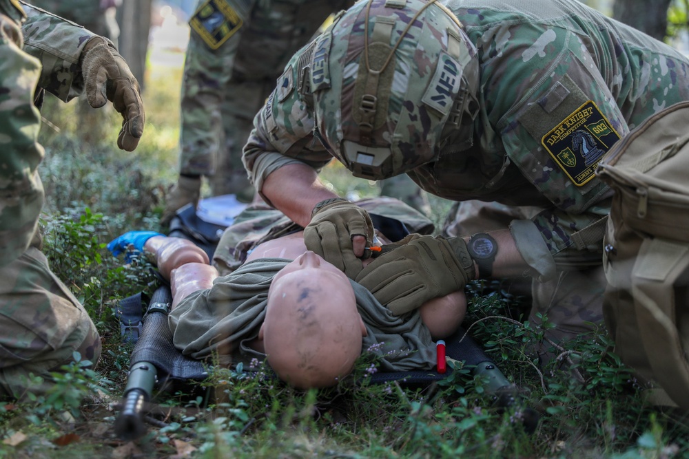 Military medics are conducting a tactical combat casualty care (TCCC) training exercise using a medical mannequin as a simulated casualty. A soldier wearing a 'MEDIC' patch and a 'Dark Knight' unit patch is performing medical assessment or airway management on the training dummy. At least two additional soldiers in MultiCam/OCP uniforms are observing or assisting with the exercise.