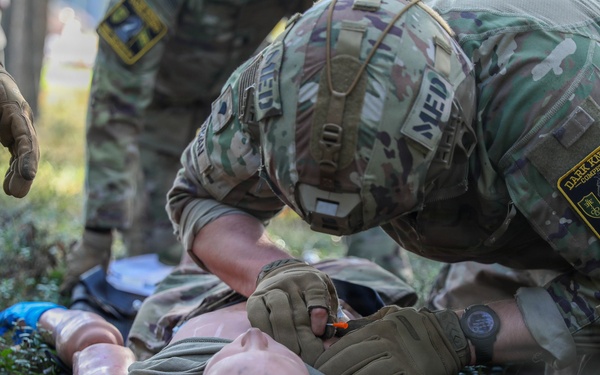 Military medics are conducting a tactical combat casualty care (TCCC) training exercise using a medical mannequin as a simulated casualty. A soldier wearing a 'MEDIC' patch and a 'Dark Knight' unit patch is performing medical assessment or airway management on the training dummy. At least two additional soldiers in MultiCam/OCP uniforms are observing or assisting with the exercise.