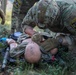 Military medics are conducting a tactical combat casualty care (TCCC) training exercise using a medical mannequin as a simulated casualty. A soldier wearing a 'MEDIC' patch and a 'Dark Knight' unit patch is performing medical assessment or airway management on the training dummy. At least two additional soldiers in MultiCam/OCP uniforms are observing or assisting with the exercise.