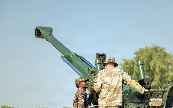 Two soldiers in different camouflage uniforms are working together on a large green artillery piece, identified by the marking 'C-0048' on its body. One soldier wears a distinctive multi-color camouflage pattern consistent with Indian Army uniforms, while the other wears a MultiCam/OCP-style pattern. The setting appears to be an outdoor training or demonstration area with trees and a low barrier wall visible in the background.