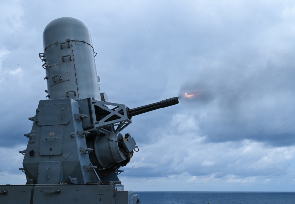 The image shows a close-up of a Phalanx Close-In Weapon System (CIWS) mounted on a naval vessel, actively firing as evidenced by the muzzle flash and smoke at the barrel tip. The weapon system is painted in standard naval gray and is photographed against an overcast sky with the ocean visible in the background. This appears to be a live-fire exercise or operational engagement at sea.