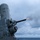 The image shows a close-up of a Phalanx Close-In Weapon System (CIWS) mounted on a naval vessel, actively firing as evidenced by the muzzle flash and smoke at the barrel tip. The weapon system is painted in standard naval gray and is photographed against an overcast sky with the ocean visible in the background. This appears to be a live-fire exercise or operational engagement at sea.