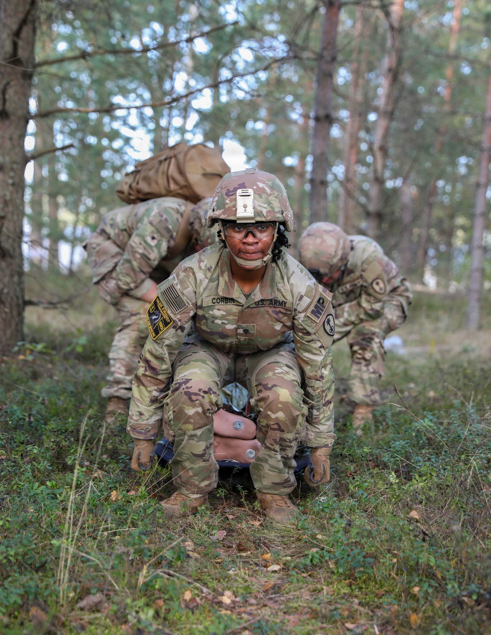 A U.S. Army soldier named Corbin, wearing OCP (Operational Camouflage Pattern) uniform with an MP (Military Police) tab, is crouched in a wooded training area while dragging or handling a simulated casualty or equipment. Two additional soldiers in the background are bent over, also engaged in the same field exercise. The setting appears to be a tactical combat casualty care (TCCC) or field training exercise in a forested environment.