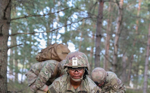 A U.S. Army soldier named Corbin, wearing OCP (Operational Camouflage Pattern) uniform with an MP (Military Police) tab, is crouched in a wooded training area while dragging or handling a simulated casualty or equipment. Two additional soldiers in the background are bent over, also engaged in the same field exercise. The setting appears to be a tactical combat casualty care (TCCC) or field training exercise in a forested environment.
