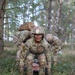 A U.S. Army soldier named Corbin, wearing OCP (Operational Camouflage Pattern) uniform with an MP (Military Police) tab, is crouched in a wooded training area while dragging or handling a simulated casualty or equipment. Two additional soldiers in the background are bent over, also engaged in the same field exercise. The setting appears to be a tactical combat casualty care (TCCC) or field training exercise in a forested environment.