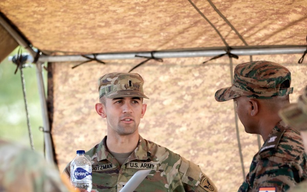 The image shows a group of military personnel from at least two different nations — the United States Army and the Indian Army — engaged in a discussion under a camouflage canopy or shade structure. A U.S. Army soldier with the name tape 'DOTZMAN' is prominently visible, holding a Kinley water bottle. This appears to be a joint military exercise or bilateral training event.
