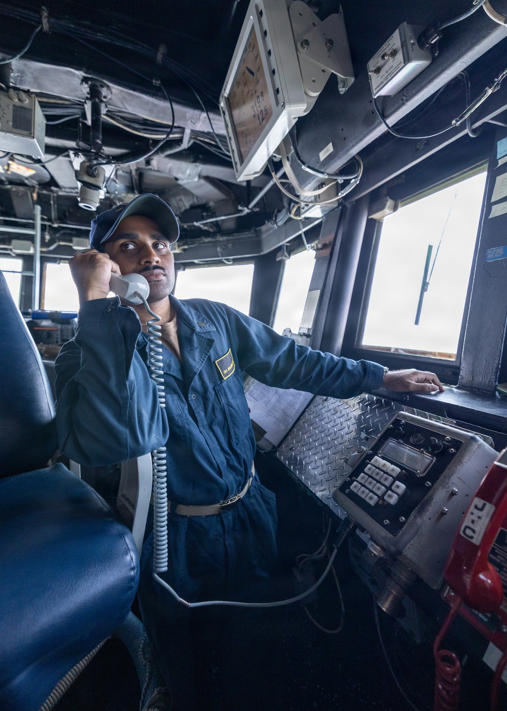 A naval or maritime crew member in blue coveralls is standing at a ship's bridge or pilothouse, holding a corded telephone handset to his ear while operating controls. He is wearing a dark cap and a name/rank patch on his chest, suggesting military or official maritime service. The environment is densely equipped with communication and navigation equipment typical of a vessel's command station.