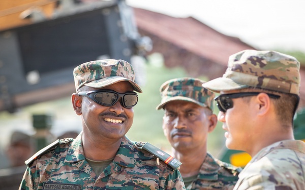 The image shows military personnel from at least two different armed forces engaged in a friendly conversation outdoors. The soldier on the left wears Indian Army digital camouflage with a visible name tape reading 'SANTOSH KORANE' and an Indian flag patch, while the soldier on the right wears US Army MultiCam (OCP) uniform with a cavalry unit patch. The interaction appears to be part of a joint military exercise or bilateral engagement.