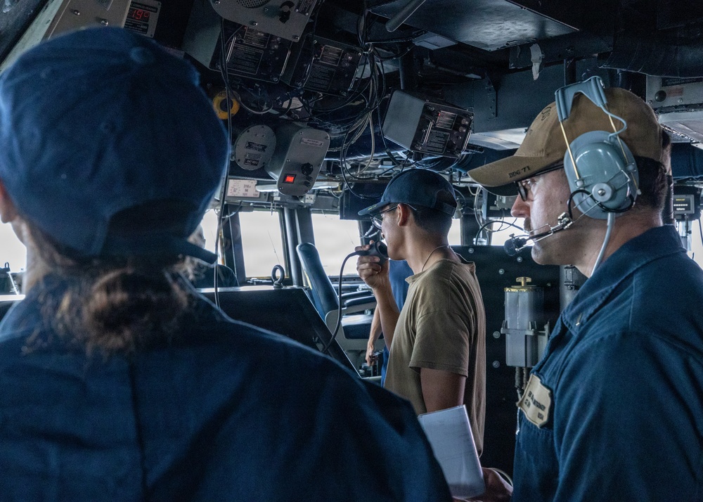 The image shows what appears to be the bridge of a U.S. Navy vessel with at least three personnel on duty. One sailor in a tan shirt is speaking into a radio handset while another in a blue uniform wears a communications headset with a microphone. The setting is clearly a ship's bridge with navigation equipment, wiring, and control panels visible overhead and around the crew.