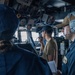The image shows what appears to be the bridge of a U.S. Navy vessel with at least three personnel on duty. One sailor in a tan shirt is speaking into a radio handset while another in a blue uniform wears a communications headset with a microphone. The setting is clearly a ship's bridge with navigation equipment, wiring, and control panels visible overhead and around the crew.