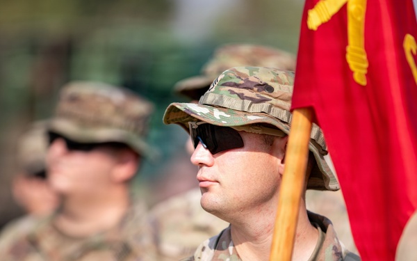 A U.S. Army soldier in Operational Camouflage Pattern (OCP) uniform stands at attention holding a red unit guidon flag with yellow insignia. The soldier is wearing tactical gear including a plate carrier vest, sunglasses, and a boonie hat. Additional soldiers in similar uniforms are visible in the blurred background, suggesting a formation or ceremony.