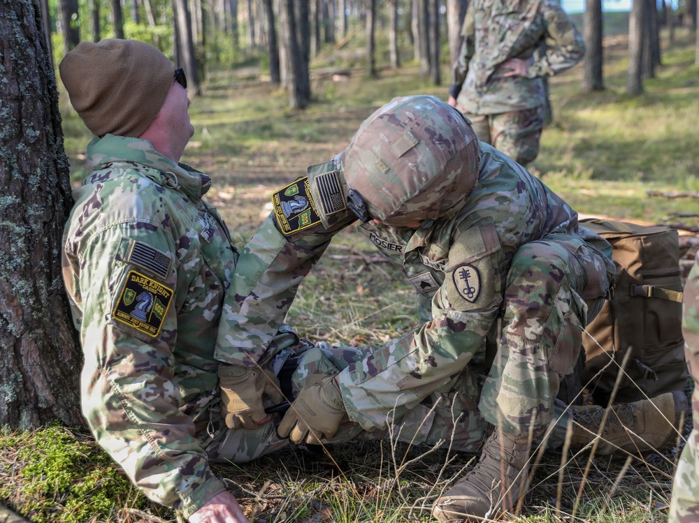 Two U.S. Army soldiers in MultiCam/OCP camouflage uniforms are engaged in what appears to be a tactical combat casualty care (TCCC) training exercise in a forested environment. One soldier, identified by name tape as 'MOSIER,' is kneeling and administering medical aid to another soldier seated against a tree. A third soldier is visible in the background observing the exercise.