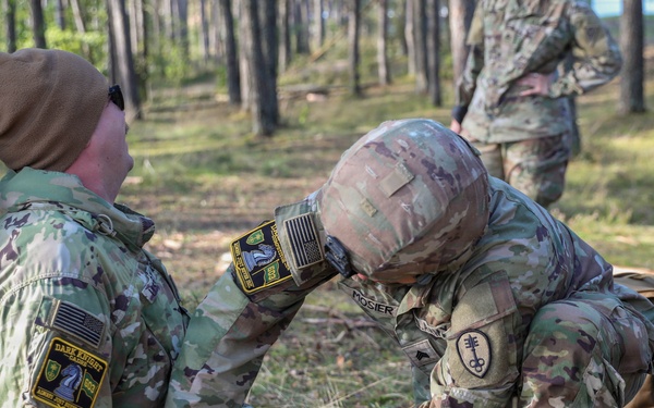 Two U.S. Army soldiers in MultiCam/OCP camouflage uniforms are engaged in what appears to be a tactical combat casualty care (TCCC) training exercise in a forested environment. One soldier, identified by name tape as 'MOSIER,' is kneeling and administering medical aid to another soldier seated against a tree. A third soldier is visible in the background observing the exercise.