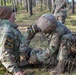 Two U.S. Army soldiers in MultiCam/OCP camouflage uniforms are engaged in what appears to be a tactical combat casualty care (TCCC) training exercise in a forested environment. One soldier, identified by name tape as 'MOSIER,' is kneeling and administering medical aid to another soldier seated against a tree. A third soldier is visible in the background observing the exercise.