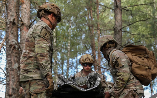 Three soldiers in US Army OCP (Operational Camouflage Pattern) uniforms are conducting a field medical evacuation exercise in a forested environment. They are carrying a litter (stretcher) with what appears to be a simulated casualty wrapped in a mylar/emergency blanket. The setting appears to be a training exercise based on the controlled environment and observable safety measures.