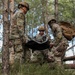 Three soldiers in US Army OCP (Operational Camouflage Pattern) uniforms are conducting a field medical evacuation exercise in a forested environment. They are carrying a litter (stretcher) with what appears to be a simulated casualty wrapped in a mylar/emergency blanket. The setting appears to be a training exercise based on the controlled environment and observable safety measures.