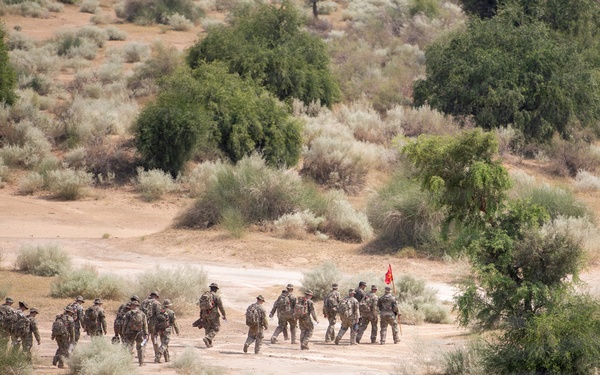 A group of approximately 20 military personnel in camouflage uniforms and full gear are conducting a foot march along a dirt trail through arid desert scrubland. One individual near the front of the group is carrying a red guidon flag, suggesting an organized unit movement or training exercise. The image is taken from an elevated vantage point, providing a wide aerial-style perspective of the formation and surrounding terrain.