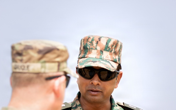 Two military personnel in different camouflage uniforms are engaged in conversation outdoors. The soldier in the foreground wears US Army MultiCam (OCP) pattern uniform, while the soldier in focus wears a digital camouflage pattern uniform with the name tape 'VINEET' visible on his chest. The soldier named Vineet is holding a handheld radio/walkie-talkie and appears to be speaking.
