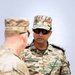 Two military personnel in different camouflage uniforms are engaged in conversation outdoors. The soldier in the foreground wears US Army MultiCam (OCP) pattern uniform, while the soldier in focus wears a digital camouflage pattern uniform with the name tape 'VINEET' visible on his chest. The soldier named Vineet is holding a handheld radio/walkie-talkie and appears to be speaking.