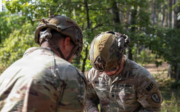 Two U.S. Army Military Police soldiers in OCP (Operational Camouflage Pattern) uniforms are working together in a wooded outdoor environment. The soldier facing the camera is identified by name tape as 'MORELAND' and wears an 'MP' branch tab with a unit patch. They appear to be handling or inspecting items near what looks like a mylar emergency blanket on the ground.