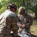 Two U.S. Army Military Police soldiers in OCP (Operational Camouflage Pattern) uniforms are working together in a wooded outdoor environment. The soldier facing the camera is identified by name tape as 'MORELAND' and wears an 'MP' branch tab with a unit patch. They appear to be handling or inspecting items near what looks like a mylar emergency blanket on the ground.