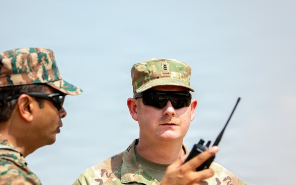 Two military personnel in different camouflage uniforms are engaged in a conversation outdoors. The soldier on the right is identified by a name tape reading 'GIAUQUE' and wears U.S. Army OCP (Operational Camouflage Pattern) uniform, while the soldier on the left wears a distinct tiger-stripe or pixelated camouflage pattern. One individual is holding a handheld radio/walkie-talkie during what appears to be a joint military exercise or coordination meeting.