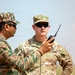 Two military personnel in different camouflage uniforms are engaged in a conversation outdoors. The soldier on the right is identified by a name tape reading 'GIAUQUE' and wears U.S. Army OCP (Operational Camouflage Pattern) uniform, while the soldier on the left wears a distinct tiger-stripe or pixelated camouflage pattern. One individual is holding a handheld radio/walkie-talkie during what appears to be a joint military exercise or coordination meeting.
