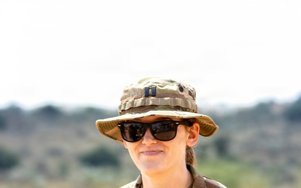 A U.S. Army soldier wearing OCP (Operational Camouflage Pattern) uniform is photographed outdoors in what appears to be an arid or semi-arid environment. The soldier is wearing a boonie hat with a Captain's rank insignia, sunglasses, and holding a helmet. The individual is smiling and appears relaxed in a field setting.