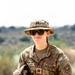 A U.S. Army soldier wearing OCP (Operational Camouflage Pattern) uniform is photographed outdoors in what appears to be an arid or semi-arid environment. The soldier is wearing a boonie hat with a Captain's rank insignia, sunglasses, and holding a helmet. The individual is smiling and appears relaxed in a field setting.
