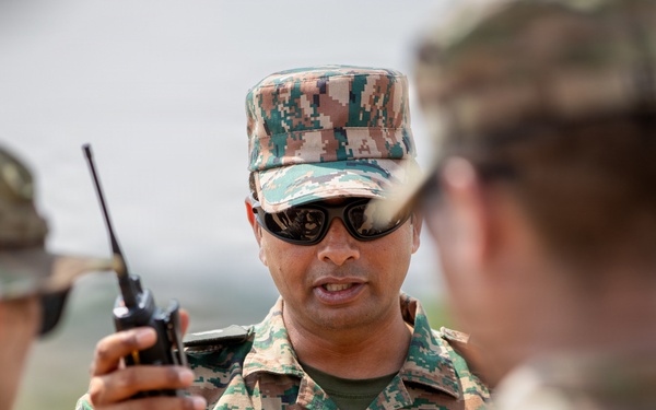 A military soldier identified by a name tape reading 'VINEET' is holding a handheld radio/walkie-talkie and appears to be briefing or communicating with other soldiers. He is wearing a distinctive digital camouflage uniform that differs from the MultiCam/OCP pattern worn by the other personnel present, suggesting a multinational or joint military exercise. The setting appears to be an outdoor training or operational environment.