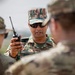 A military soldier identified by a name tape reading 'VINEET' is holding a handheld radio/walkie-talkie and appears to be briefing or communicating with other soldiers. He is wearing a distinctive digital camouflage uniform that differs from the MultiCam/OCP pattern worn by the other personnel present, suggesting a multinational or joint military exercise. The setting appears to be an outdoor training or operational environment.
