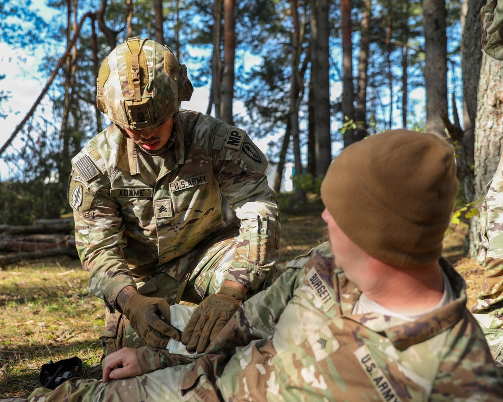 A U.S. Army Sergeant named Adame, wearing OCP camouflage and a tactical helmet with an 'IMP' patch, is performing what appears to be a medical treatment or first aid procedure on another soldier named Burgett in a forested outdoor setting. The soldier receiving treatment is seated on the ground wearing a brown beanie cap. This appears to be a training exercise based on the controlled environment.