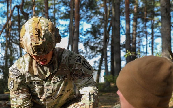 A U.S. Army Sergeant named Adame, wearing OCP camouflage and a tactical helmet with an 'IMP' patch, is performing what appears to be a medical treatment or first aid procedure on another soldier named Burgett in a forested outdoor setting. The soldier receiving treatment is seated on the ground wearing a brown beanie cap. This appears to be a training exercise based on the controlled environment.