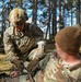 A U.S. Army Sergeant named Adame, wearing OCP camouflage and a tactical helmet with an 'IMP' patch, is performing what appears to be a medical treatment or first aid procedure on another soldier named Burgett in a forested outdoor setting. The soldier receiving treatment is seated on the ground wearing a brown beanie cap. This appears to be a training exercise based on the controlled environment.