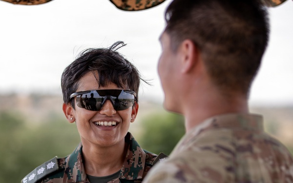 Two military personnel are engaged in a friendly conversation outdoors. The individual facing the camera is wearing Indian Army digital camouflage uniform with a name tape reading 'AISHWARYA KHORATE' and rank insignia visible on the shoulders. The second individual, seen from behind, is wearing a MultiCam/OCP pattern uniform consistent with U.S. military dress.
