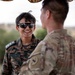 Two military personnel are engaged in a friendly conversation outdoors. The individual facing the camera is wearing Indian Army digital camouflage uniform with a name tape reading 'AISHWARYA KHORATE' and rank insignia visible on the shoulders. The second individual, seen from behind, is wearing a MultiCam/OCP pattern uniform consistent with U.S. military dress.