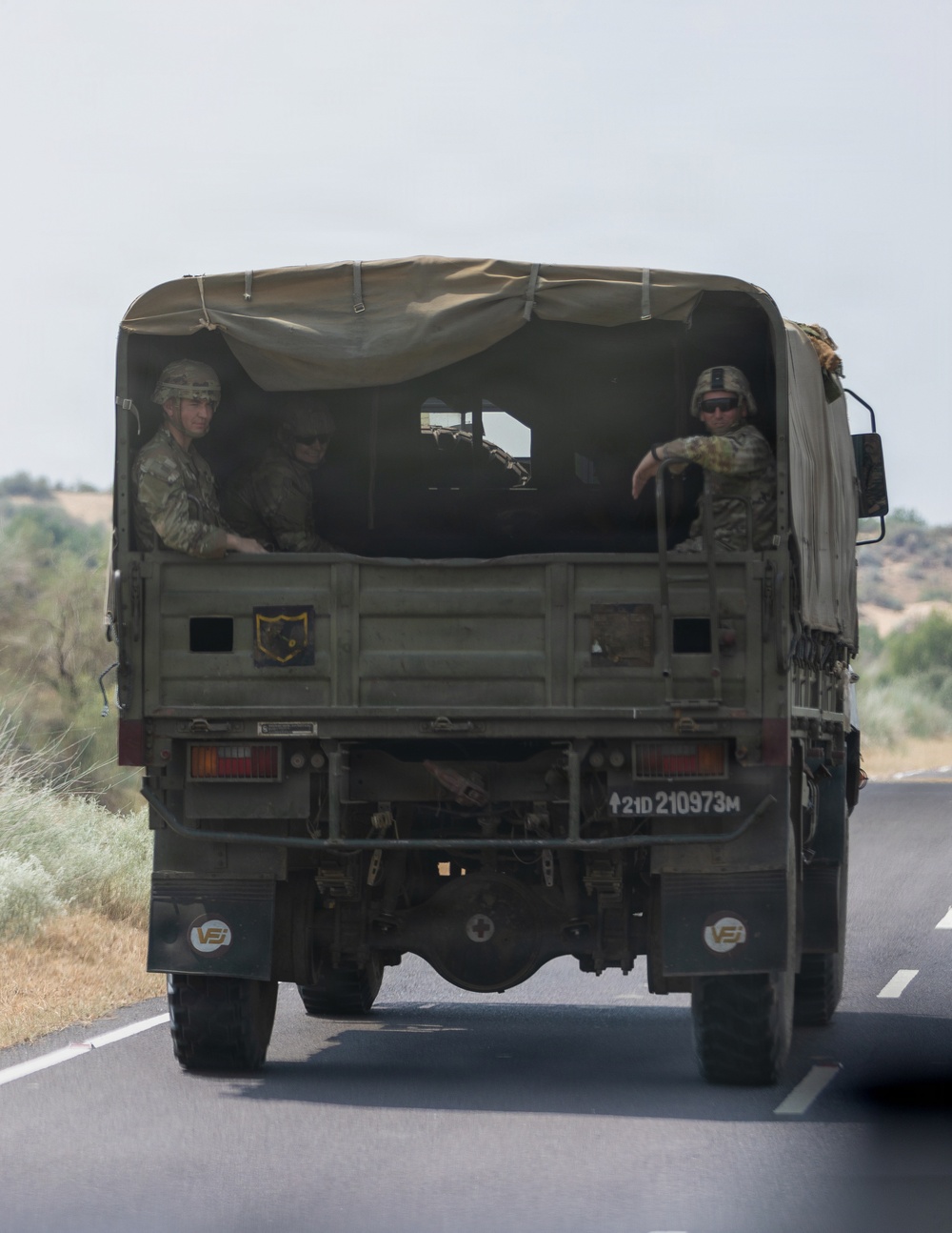 A military cargo truck is traveling on a paved road, viewed from behind. The truck carries several soldiers in camouflage uniforms and helmets in the open cargo bed. A unit insignia patch and registration number '21D 210973M' are visible on the rear of the vehicle.
