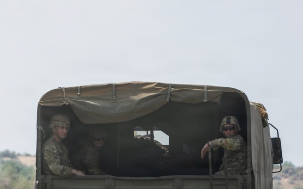 A military cargo truck is traveling on a paved road, viewed from behind. The truck carries several soldiers in camouflage uniforms and helmets in the open cargo bed. A unit insignia patch and registration number '21D 210973M' are visible on the rear of the vehicle.