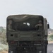 A military cargo truck is traveling on a paved road, viewed from behind. The truck carries several soldiers in camouflage uniforms and helmets in the open cargo bed. A unit insignia patch and registration number '21D 210973M' are visible on the rear of the vehicle.
