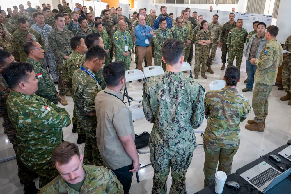 A large multinational military briefing or conference is taking place indoors, with personnel from multiple nations gathered in a semicircle around a speaker. Participants are wearing various camouflage uniforms representing different countries, including what appear to be Singapore, Australian, and US military forces, along with several civilians in business casual attire. The setting appears to be a formal military exercise or joint training event, with presentation boards visible in the background.