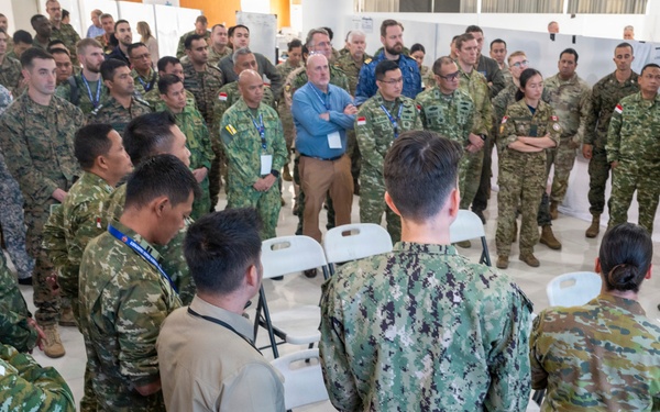 A large multinational military briefing or conference is taking place indoors, with personnel from multiple nations gathered in a semicircle around a speaker. Participants are wearing various camouflage uniforms representing different countries, including what appear to be Singapore, Australian, and US military forces, along with several civilians in business casual attire. The setting appears to be a formal military exercise or joint training event, with presentation boards visible in the background.