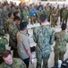 A large multinational military briefing or conference is taking place indoors, with personnel from multiple nations gathered in a semicircle around a speaker. Participants are wearing various camouflage uniforms representing different countries, including what appear to be Singapore, Australian, and US military forces, along with several civilians in business casual attire. The setting appears to be a formal military exercise or joint training event, with presentation boards visible in the background.