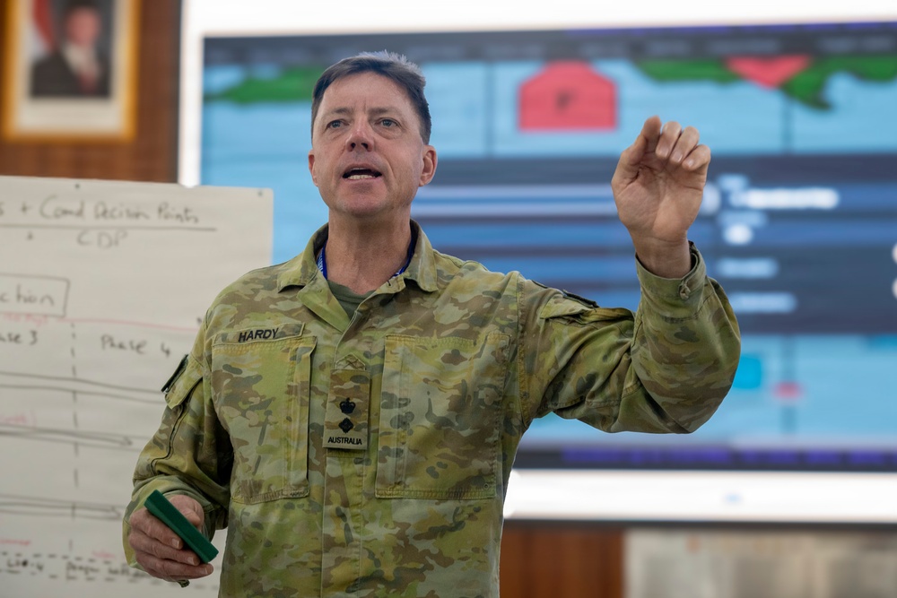 An Australian military officer named Hardy is delivering a briefing or presentation in what appears to be a command or operations room. He is gesturing with his right hand while holding a marker in his left, standing in front of a whiteboard and a large display screen showing a map or operational graphic. The setting suggests a military planning or training exercise environment.